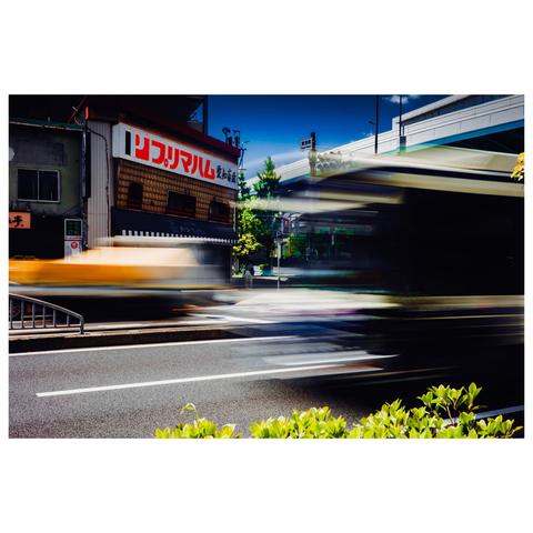 The foreground is consumed by streaks of motion: a city bus rushes from right to left, its form nearly transparent, while a truck draped in a yellow tarpaulin cuts through in the opposite direction.