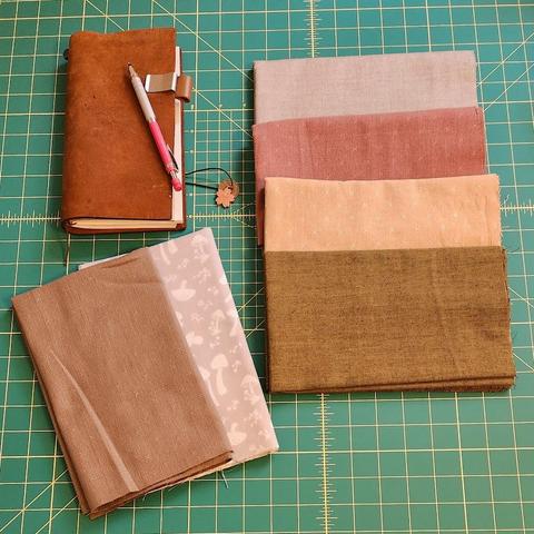Fabric is arranged on a cutting mat next to a leather traveler's notebook and mechanical pencil.
The four main colors are grey, pink, tan, and green. Where the pink and tan have small hatches in them for texture. To the side is a grey fabric with white mushrooms and a muted brown with small hatches.
