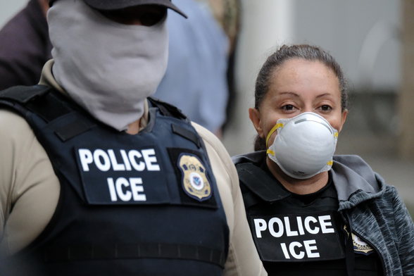 Photo of a male ICE agent wearing both a badge AND a ‘gator’ mask that hides his entire face. Interestingly, next to him is a female (apparently Hispanic) ICE agent wearing a medical mask, but whose eyes appear to be questioning her participation.