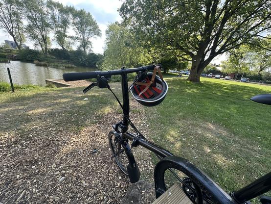 a black bicycle parked by the pond, with a helmet hanging from the handlebars and a tree-lined path and parked cars in the background.