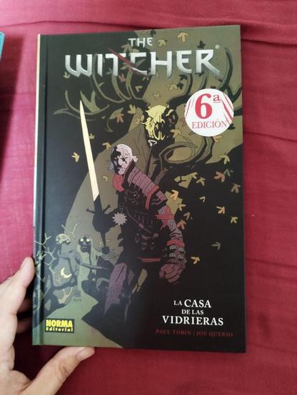 Portada del cómic 'The Witcher: La Casa de las Vidrieras' escrito por Paul Tobin e ilustrado por Joe Querio. La ilustración muestra a un personaje sosteniendo una espada, rodeado de diversas criaturas y follaje. En la portada hay una etiqueta que indica que esta es la sexta edición. En la esquina inferior derecha aparece el logo de la editorial NORMA Editorial.