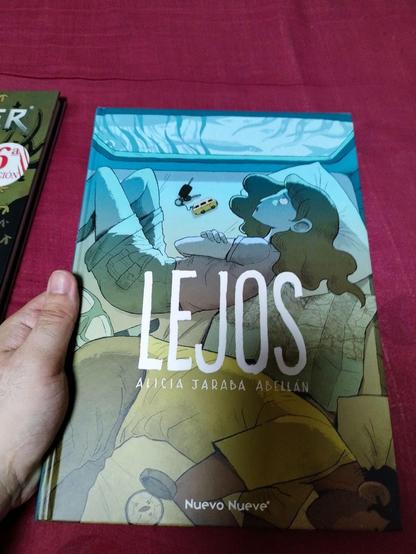 Portada del libro 'LEJOS' de Alicia Jaraba Abellán. La ilustración de la cubierta muestra a una chica con el cabello largo sentada en una cama, mirando a través de una ventana. Junto a ella hay un coche de juguete y una llave sobre la cama. En el fondo, hay un chico acostado en la cama, de espaldas a la chica. El libro está publicado por 'Nuevo Nueve'