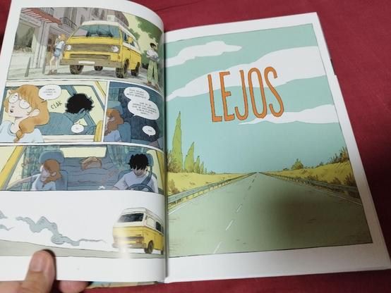 Doble página de un cómic abierto. La página izquierda contiene cuatro viñetas: la primera muestra una furgoneta amarilla alejándose con humo saliendo del escape; la segunda revela el interior del vehículo con dos personajes sentados en los asientos delanteros, uno de ellos con gafas y cabello castaño, el otro con cabello negro; la tercera muestra la furgoneta estacionada frente a un edificio; y la cuarta presenta un diálogo entre los personajes en español. La página derecha exhibe una ilustración de una carretera vacía que se extiende hacia el horizonte, con un cielo azul y nubes blancas, y la palabra 'LEJOS' escrita en grandes letras naranjas.