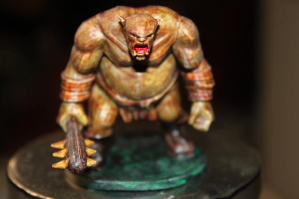 Alt Text (Front View):
A painted miniature of a hulking ogre-like creature with mottled yellow-brown skin and a furious red-mouthed expression. It holds a large spiked club in its right hand and wears leather bracers and a loincloth. The figure stands on a round green base.

Alt Text (Rear View):
Rear view of the same ogre miniature, showing its broad muscular back, leather loincloth, and bracers. The figure's posture is hunched forward, with the spiked club lowered behind it, all mounted on a green circular base.