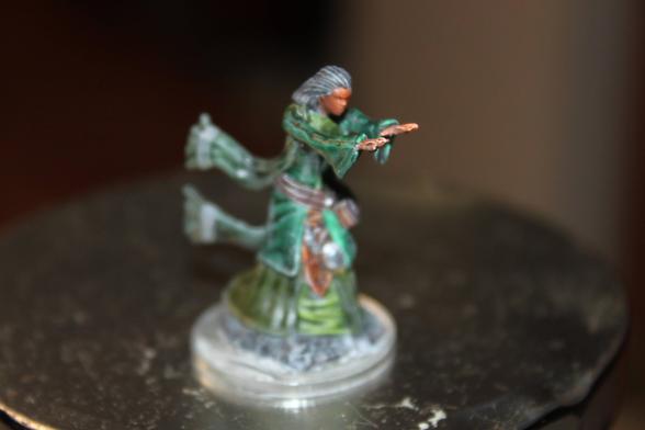 Alt Text:
A painted miniature of a spellcaster in motion, featuring a figure with dark skin and flowing gray hair, wearing green robes with light accents and a brown belt. The character is posed with both arms extended forward, as if casting a spell, and stands on a clear round base.