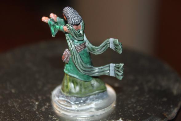 Alt Text:
Rear view of a painted miniature depicting a spellcaster in a flowing green robe with trailing sleeves, posed mid-cast with both arms extended forward. The figure has long gray hair and a belt with pouches, standing on a clear round base.