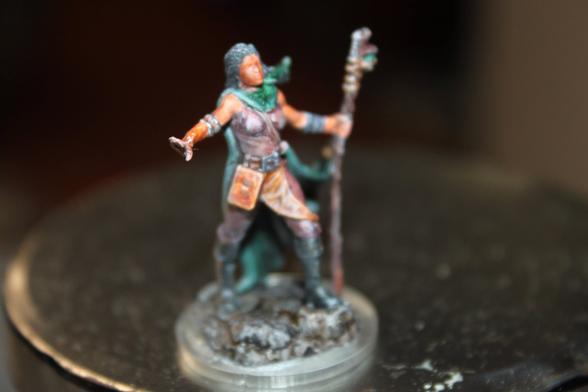 Alt Text:
A painted miniature of a female adventurer with dark hair, wearing a green scarf and  leather armor. She holds a staff in one hand and extends the other arm outward in a commanding pose. The figure stands on a textured base resembling rocky terrain.