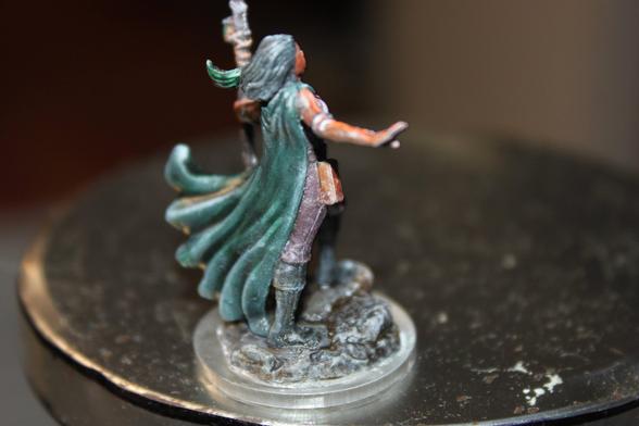 Alt Text:
Rear view of a painted miniature of a female spellcaster. She has dark hair and wears a flowing green cape, leather pants, and a green scarf. She holds a staff in one hand and gestures with the other, standing on a textured rocky base.