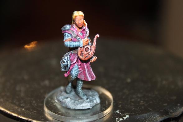 Alt Text:
Painted miniature of a bard holding a lyre. The character has light blonde hair and wears a purple tunic with metallic armor accents and a satchel at their side. They stand on a textured rocky base, with one hand poised mid-performance.