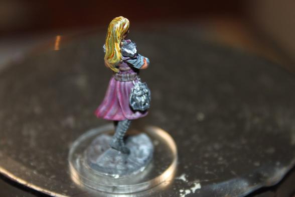 Alt Text:
Rear view of a painted bard miniature with long blonde hair wearing a purple tunic and silver shoulder armor. A detailed satchel hangs at their side, and they stand on a textured rocky base with their arms crossed.