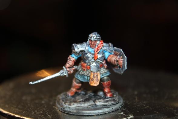 Alt Text:
A painted dwarf warrior miniature in full plate armor with a red braided beard and blue undershirt, holding a sword in one hand and a shield in the other. The figure stands in a battle-ready stance on a textured rocky base.