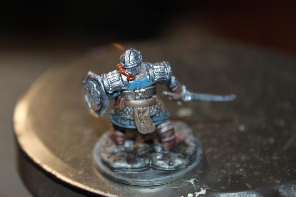 Alt Text:
A rear view of a painted dwarf warrior miniature clad in silver plate armor with a blue undershirt, red braided beard, and brown boots. The figure holds a round shield in one hand and a sword in the other, posed on a rocky base.
