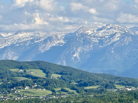 A scenic view of snow-capped mountains in the background, with lush green hills and scattered small houses in the foreground under a partly cloudy sky.