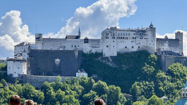 A large, historic castle sits atop a hill, surrounded by greenery and under a blue sky with scattered clouds. The castle features various towers and walls, showcasing a blend of architectural styles. In the foreground, two people are partially visible.