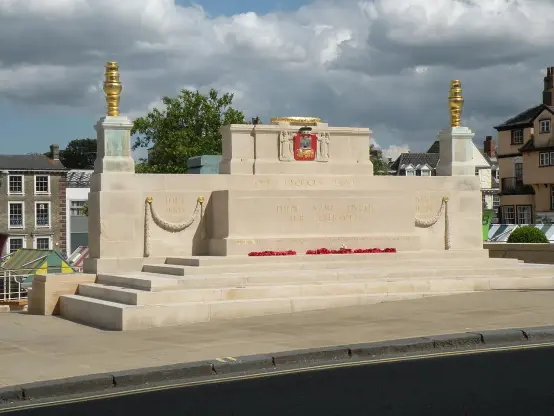 The image depicts a war memorial, likely dedicated to soldiers who fought in a particular conflict, as suggested by the dates "1914" and "1918," which correspond to World War I. The memorial features a raised stone structure with decorative elements, including a crest or emblem at the center and two torches or lamps on either side. There are also red poppies, a common symbol of remembrance for fallen soldiers, placed at the base of the memorial. Surrounding the memorial, there seems to

Image Credits: Wikimedia / Robert Edwards / CC BY-SA 2.0