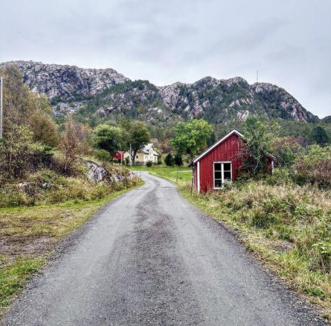 This image features a rural landscape dominated by lush greenery and a rugged mountain background. A gravel road curves through the scene, leading past a small, traditional red wooden shed on the left and onward to a cluster of houses. There's a two-story yellow house visible in the distance that adds a quaint, residential feel to the otherwise natural setting. The overall vibe is serene and picturesque, typical of Scandinavian countryside environments.