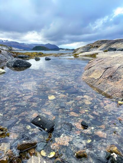 The image shows a clear and shallow body of water with a rocky bottom visible through it. An old camera is submerged in the water, near the center of the image, partially covered by algae or similar aquatic plants. The surrounding landscape features smooth, rounded rocks, while in the distance, a rugged mountainous terrain and overcast sky can be seen. The atmosphere is serene and slightly melancholic, emphasized by the moody sky and the abandoned camera in the water.