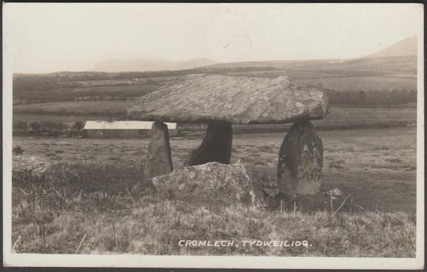 A vintage photographic postcard of the c.1910s showing a side view of the Mynydd Cefn Amlwch Dolmen or Coetan Arthur at Tydweiliog or Tudweiliog
on the Lleyn Peninsula in North Wales. The large capstone sits on three uprights, with a few other stones on the deck. The Lleyn Peninsula can be seen in the distance, beyond a long low shed in the middle distance. The dolmen is now fenced off quite tightly.