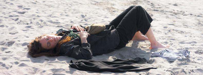 The image shows a person lying on a sandy surface, possibly a beach. They are dressed in long dark clothing and appear to be resting or asleep. There are additional clothing items, like a striped garment, on the sand next to them.