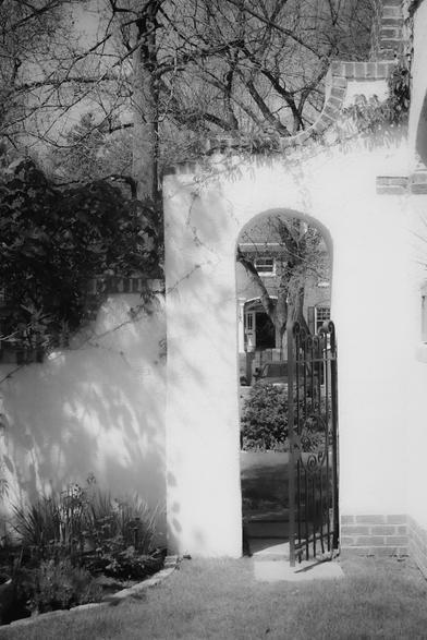 The image depicts a black and white scene featuring an arched gateway with a wrought iron gate. The gateway is part of a white wall with brick accents at the base. The gate is open, revealing a view of a garden and a house in the background. The garden includes various plants and shrubs, with a tree visible to the left of the arch. The house has a brick facade and a visible window. The scene is framed by bare tree branches, suggesting a winter or early spring setting. The sunlight casts shadows on the wall, adding depth to the image.

Provided by @altbot, generated privately and locally using Ovis2-8B