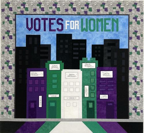 Quilt top featuring a city skyline representing when different racial categories of women won the right to vote in the U.S., displayed in full on a design wall. There are five scrappily pieced unique buildings in the foreground each with a machine embroidered sign representing racial class and an address representing the year; from left to right they are 

White Women/1920 (in purples)
Asian Women/1952 (in greens)
Native Women/1963 (very light greyish white)
Black Women/1965 (greens)
Latina Women/1975 (purples)

Behind the these buildings, which resemble a votes for women flag, are scrappily pieced black buildings set against a bright blue sky where the words VOTES FOR WOMEN have been pieced from purple, white and green fabrics. Surrounding this center section on both sides and the top is a small black and grey stripe border and a wider border of a black and white skyline print that has had random groupings of buildings painted in purple and green. The bottom border consists of a small light grey strip resembling a sidewalk and a wider section of mirrored black, purple, green and white fabrics that slant outward like rays.