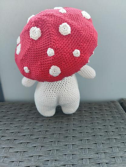 A crocheted amigurumi toy resembling a mushroom, handcrafted with white cotton yarn for the body and red yarn with white spots for the cap. The plush mushroom sits on a woven surface with a simple, neutral background.