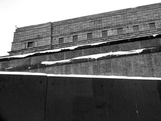 a black and white photograph of the Finnish Parliament in Helsinki, a large, imposing stone building with a blocky, institutional architecture. The structure features a stone brick facade with rows of small, barred or gridded windows visible along its upper section. There are several levels of stone walls or terracing in the foreground, with patches of snow visible along the edges. The stark, monumental style and heavy masonry creates a severe, austere appearance against a plain sky background. The composition emphasizes the building's massive scale and geometric forms through the upward perspective. (made with Claude.ai)