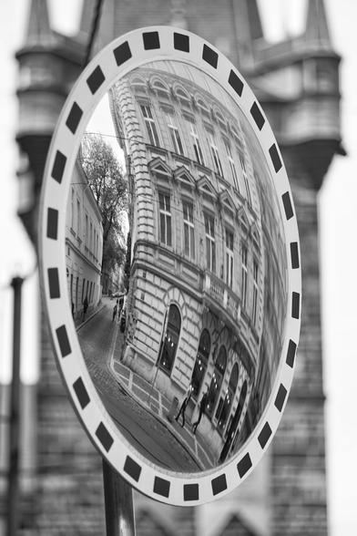 Schwarz-Weiß-Foto im Hochformat. Im Hintergrund unscharf ein Turm, im Vordergrund ein Verkehrsspiegel, in dem sich ein Prager Altbau spiegelt.