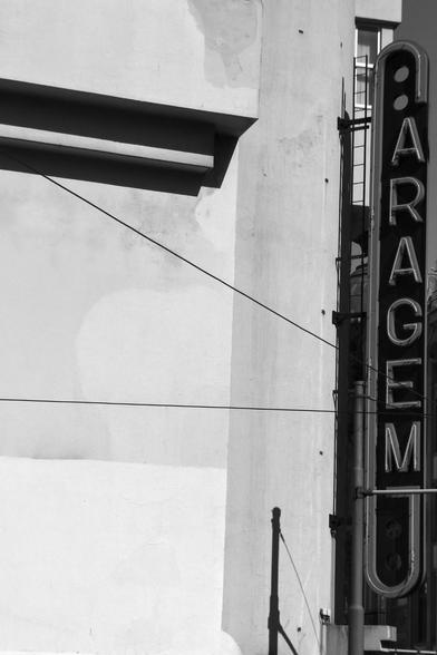 Black and white photo of a wall with some strong lines and shadows in black against a pale background. To the right the wall ends in a vertical neon sign which reads 'Garagem'. The initial G is extremely elongated so that all the other letters are stacked within its upper and lower jaws.