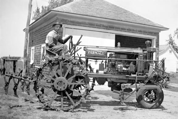 A man in overalls and long gloves riding a 'Farmall' tractor. The tractor has no outer shell, so the engine and other parts are visible. In the background is a brick garage with a shingle roof and a sliding, barn-style door.