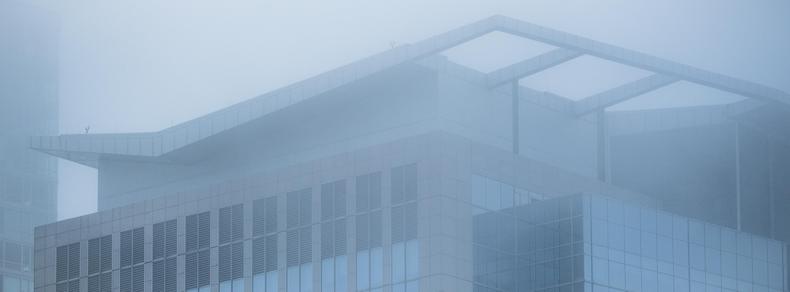 An urban building – in shades of mid-blue, dark blue and gray – is rendered in softened contrast courtesy of the low-flying cloud which engulfs its top.  The building wears a fascinator shaped like a zippy flattened "V", which feature is especially befogged.