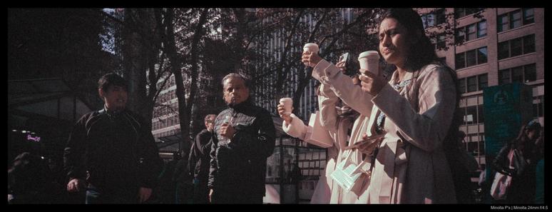 Three people in coats hold up coffees with the intent of snapping photos with their phones.  A few bystanders look at them.