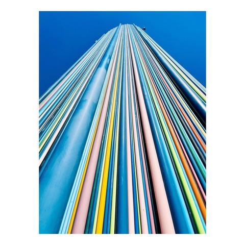 A vertical view of colorful striped columns against a clear blue sky. The columns feature various shades, including blue, pink, and yellow, creating a vibrant pattern.