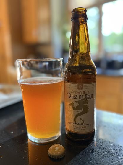 A pint glass of golden honey colored ale. The brown bottle is to the right with a white and gold label and a dragon with furled wings in silhouette