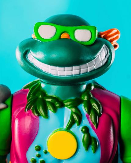 Close-up of a Teenage Mutant Ninja Turtle action figure wearing green sunglasses, a pink vest, and a leafy necklace, set against a blue background.