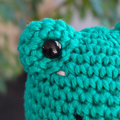 A close up of a crocheted frog face with embroidered pink cheeks.