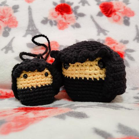 - white blanket with patterns: grey Eiffel Towers, red/pink roses with grey leaves
- centered: two crochet amigurumi dolls of different sizes
- both have black hair with bangs, two black bead eyes, beige skin, black bottom (clothing)
- the smaller doll has a black crocheted looped string on their head so it can be easily carried around