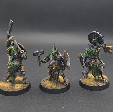 3 Beastmen wearing green hoods. Left, has a 2 handed axe and a shield on it's back. Centre has a hammer and shield (the shield has two heads attaches to the outside) and right, has a shield held high and a mace