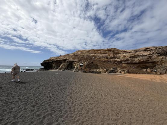 Blick über einen Sandstrand mit nicht ganz hellem Sand auf eine Felsformation in hellem STein. Der Himmel.
