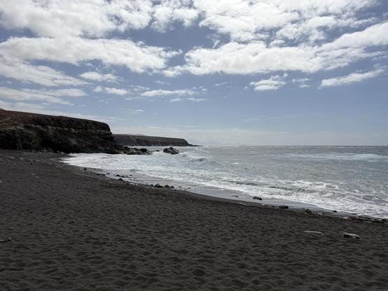 Blick über eine Strand mit dunklem Sand. Die Aufnahme ist etwas im Gegenlict so dass der Sand sehr dunkel erscheint, aber das Licht auf den flachen Wellen am Strand glitzert. in der Ferne sind steile Klippen zu sehen .Der Himmel ist leicht bewölkt