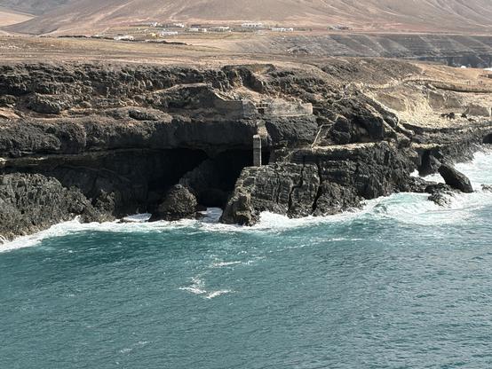 Bild aus erhöhter Postition auf eine Bucht in der zerklüftete Klippen aus dunklem STein zu sehen sind Es gibt dort dunkle Vertiefungen, deren Tiefe man nicht einsehen kann. Das türkisfarbene Meer schlägt mit weißer Gischt an die Klippen