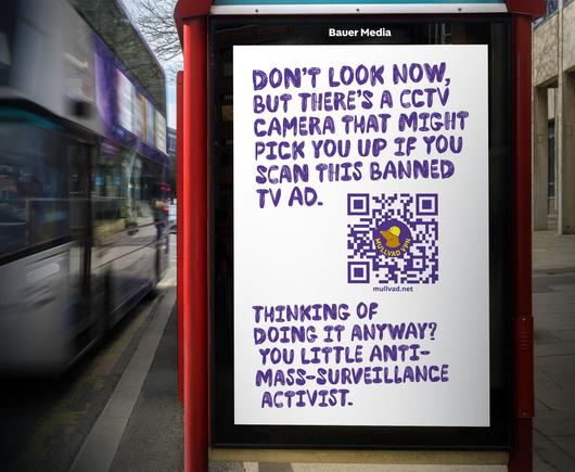 An image of the advert on the side of a bus stop.

The background is white, with purple stylised text which reads: "Don't look now, but there's a CCTV camera that might pick you up if you scan this banned TV ad."

Below is a QR code which leads to mullvad.net, the QR code is purple, and has the Mullvad VPN logo and mole inside.

Underneath is more text which reads: "Thinking of doing it anyway? You little anti-mass-surveillance activist."