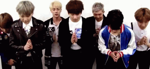 Seven young Korean men dressed in casual attire, some with dyed gray, blonde, and dark red hair bowing with their hands together.