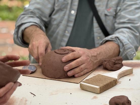 Two hands working with brown clay on a table. The maker cups an upside down bowl with one hand and uses his other to shape the object. The bow has a small foot ring. On the table nearby is a paddle tool, lumps of clay and another participants hands molding clay.