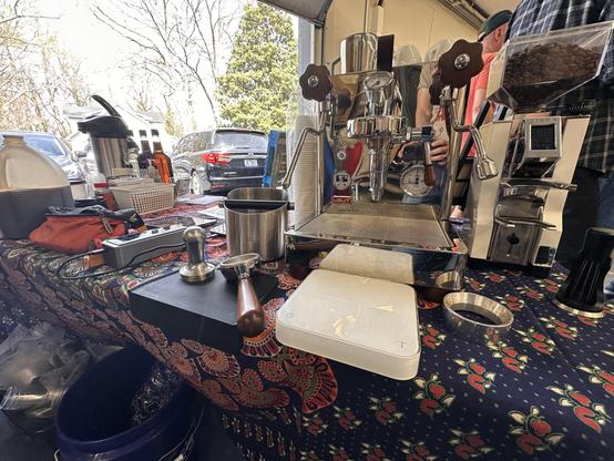 Photo of a chrome espresso machine with a white espresso machine next to it on a table where there are other coffee supplies