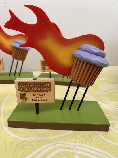 A trophy that is handmade out of wood, and is a cupcake with purple frosting with red and yellow flames coming out of the back of it. This award is for the  Anti-Fascist Cake Contest Winner Best Pun'd Cake