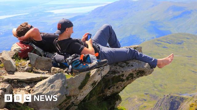Two male walkers resting on rock overhang on top of yr Wyddfa in north Wales. There is hiking equipment besides them including some walking shoes.