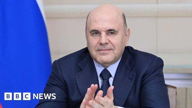 Russian Prime Minister Mikhail Mishustin claps his hands at a ceremony. The blue of a Russian flag falls behind his left shoulder.