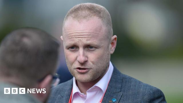 Jamie Bryson has blond short hair, shaved in a number one cut.  He is wearing a grey suit and pink shirt.  He is talking to a man whose head is in shot from behind. 