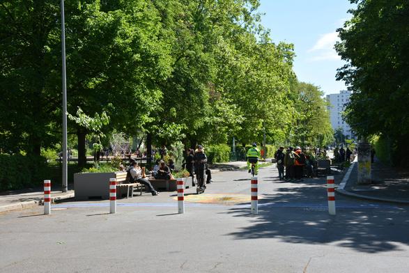 Die Schulstraße in Berlin-Mitte, Singerstraße. Kinder und Jugendliche sitzen auf Bänken, eine Person fährt auf dem E-Roller, eine andere auf dem Rad. Es ist sonnig und sehr grün.